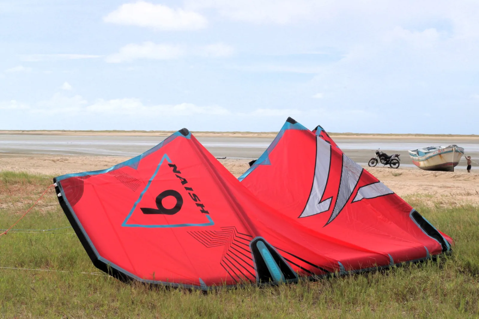 Kite inflável vermelho da Naish aberto no chão gramado da Ilha do Guajirú com mar ao fundo